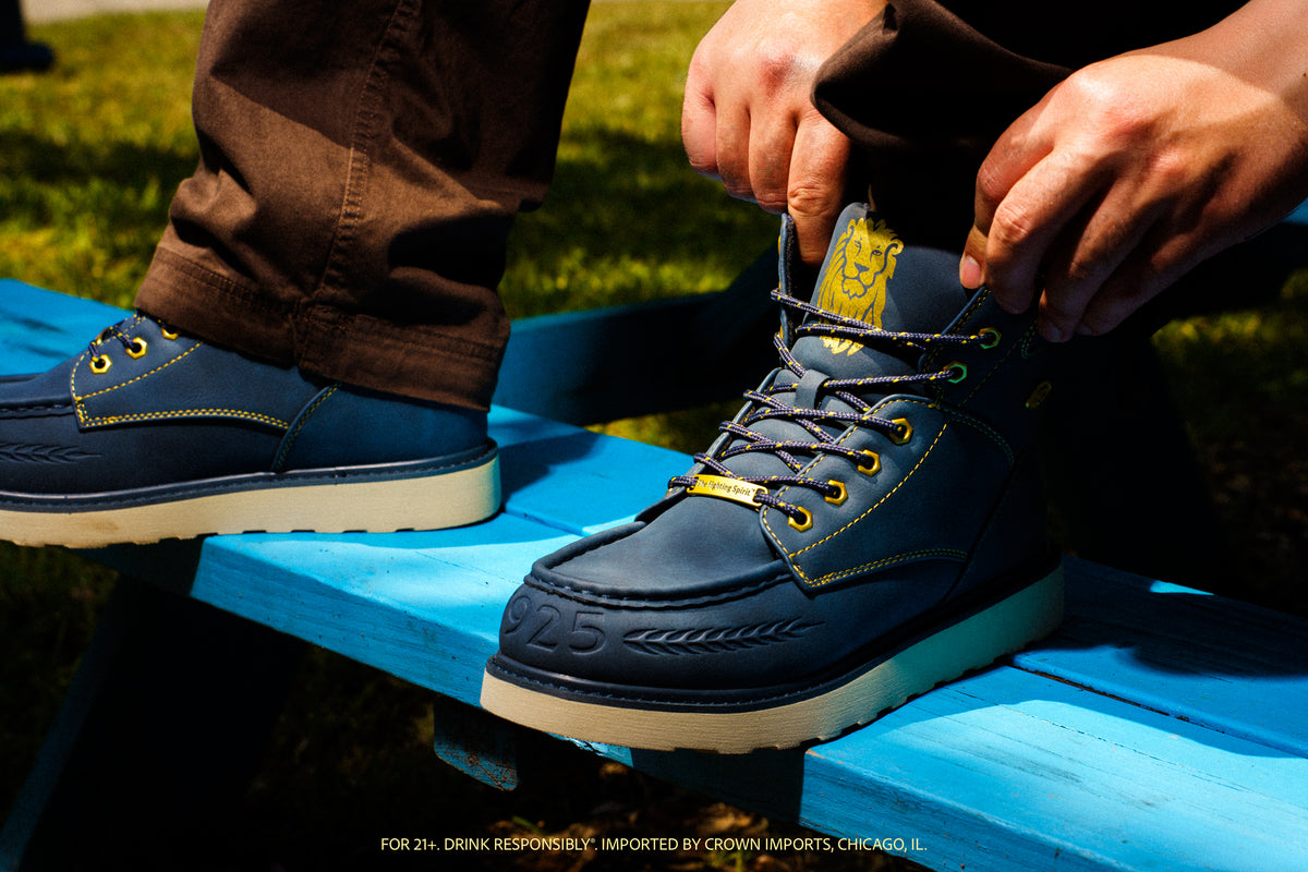 A person sitting outdoors on a blue bench putting on a dark navy blue boot. The boot is a collaboration with Modelo and has gold stitching and co-branded details on a durable wedge sole