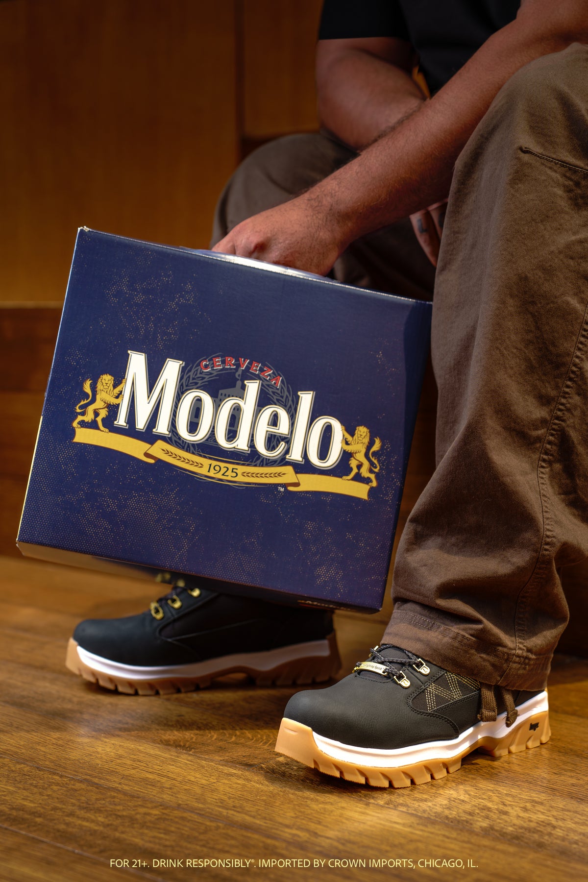 A person sitting on a wooden floor holds a large blue Modelo shoe box while wearing the Wilderness boot from the collection. The boot is black with gold stitching and gum soles. The Modelo logo is clearly visible on the box.