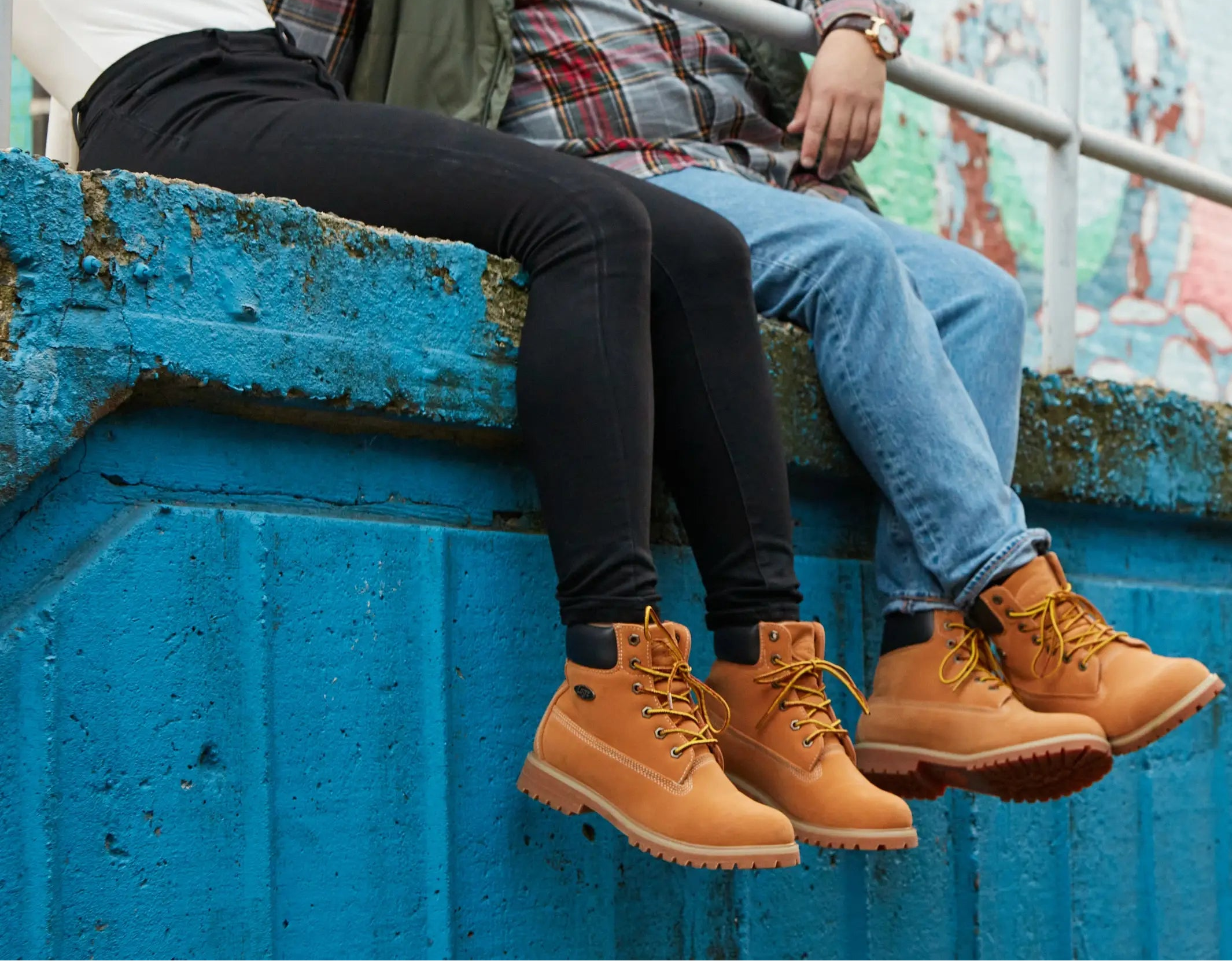 Two people sitting on a blue wall wearing brown boots with a colorful mural in the background.