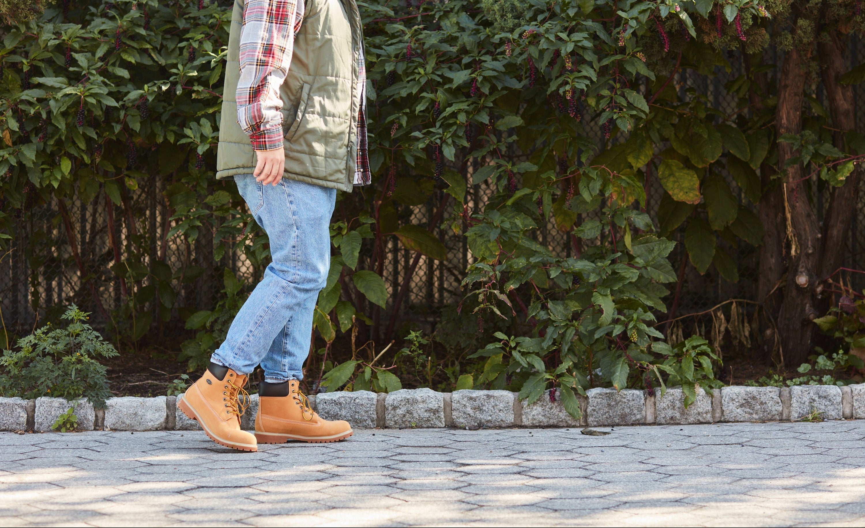 Person wearing a red beanie, plaid shirt, green vest, blue jeans, and brown boots walking outdoors.
