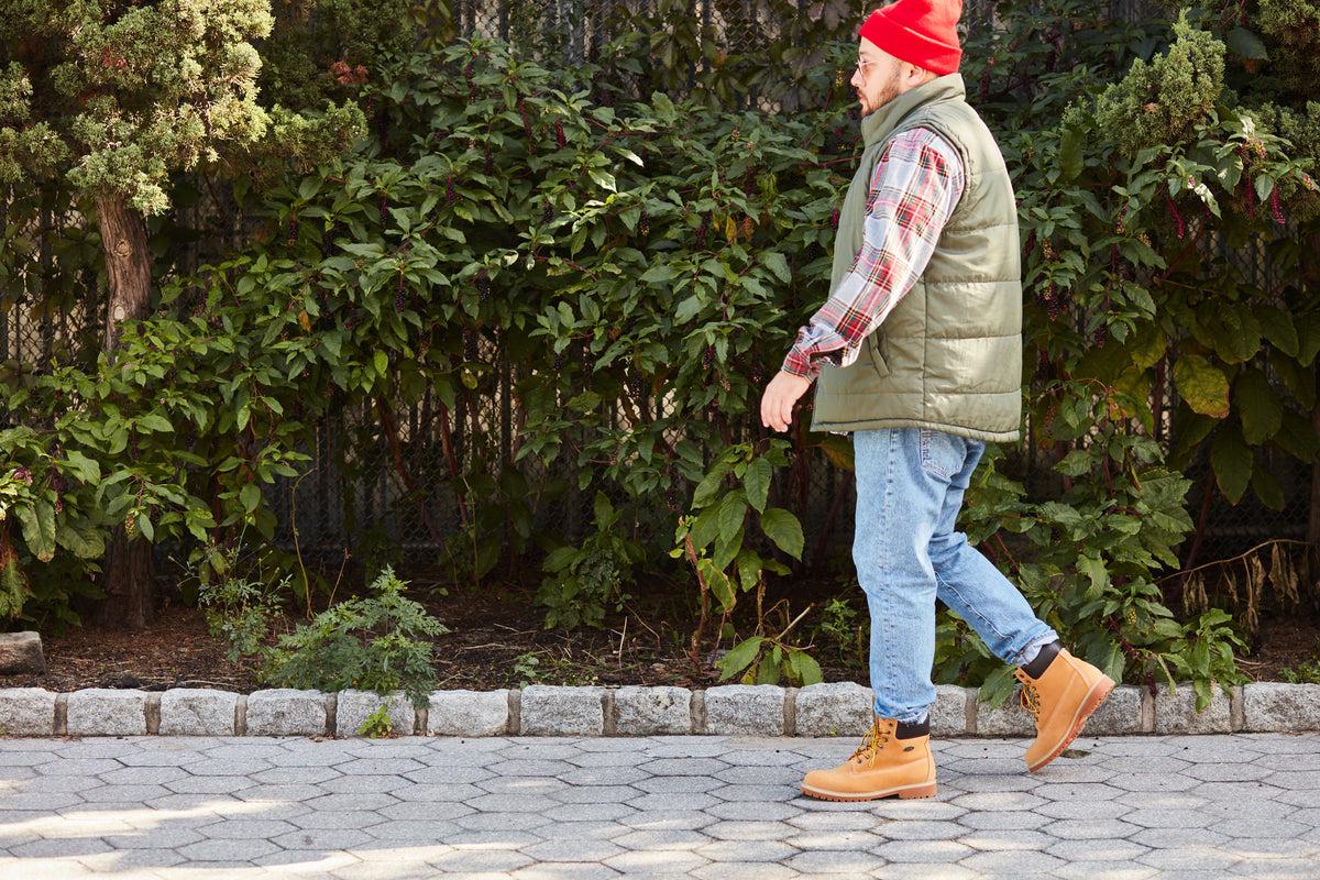 A man walking outdoors on a paved path wearing a red knit beanie, an olive green quilted puffer vest over a red and white plaid flannel shirt, light blue jeans, and Lugz Convoy boots, with a green garden background.
