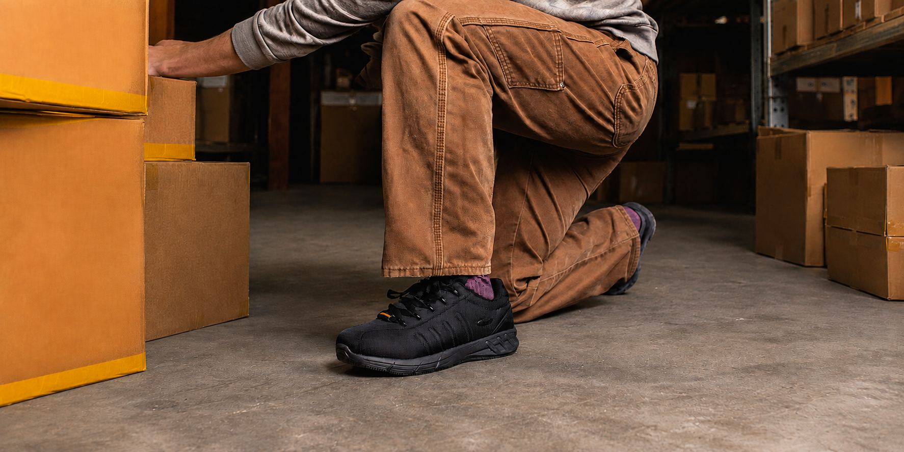 A person kneeling in a warehouse next to cardboard boxes, wearing brown work pants, a gray long-sleeve shirt, and black work shoes with purple socks visible.
