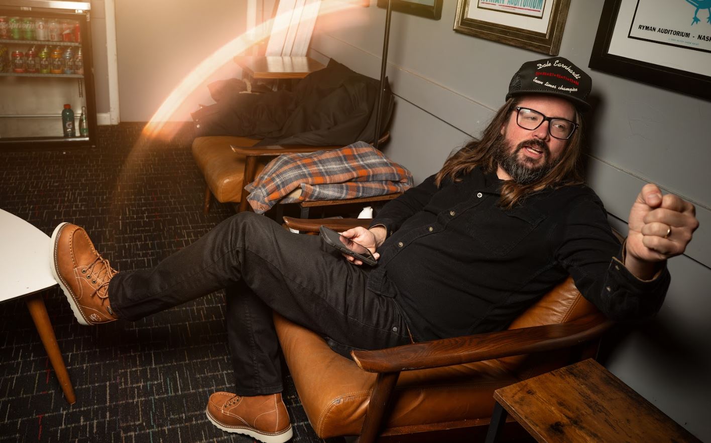 Man sitting in a chair in a cozy room with a plaid blanket and framed pictures on the wall.