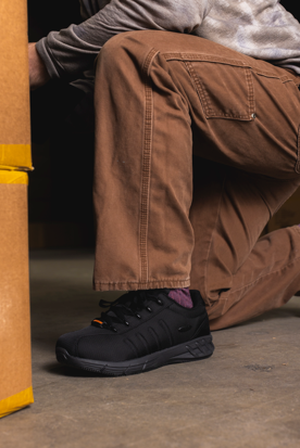 A close-up of a person wearing black work boots with brown cargo pants, kneeling on a concrete floor next to a cardboard box.