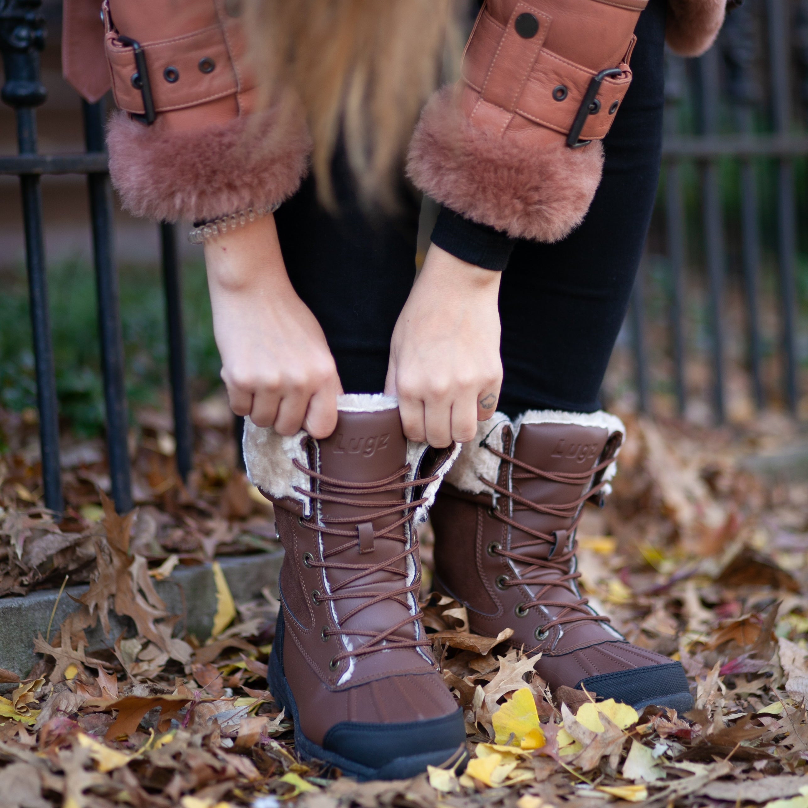 Person wearing brown winter boots with fur-lined hood and cuffs, standing on a leaf-covered ground.